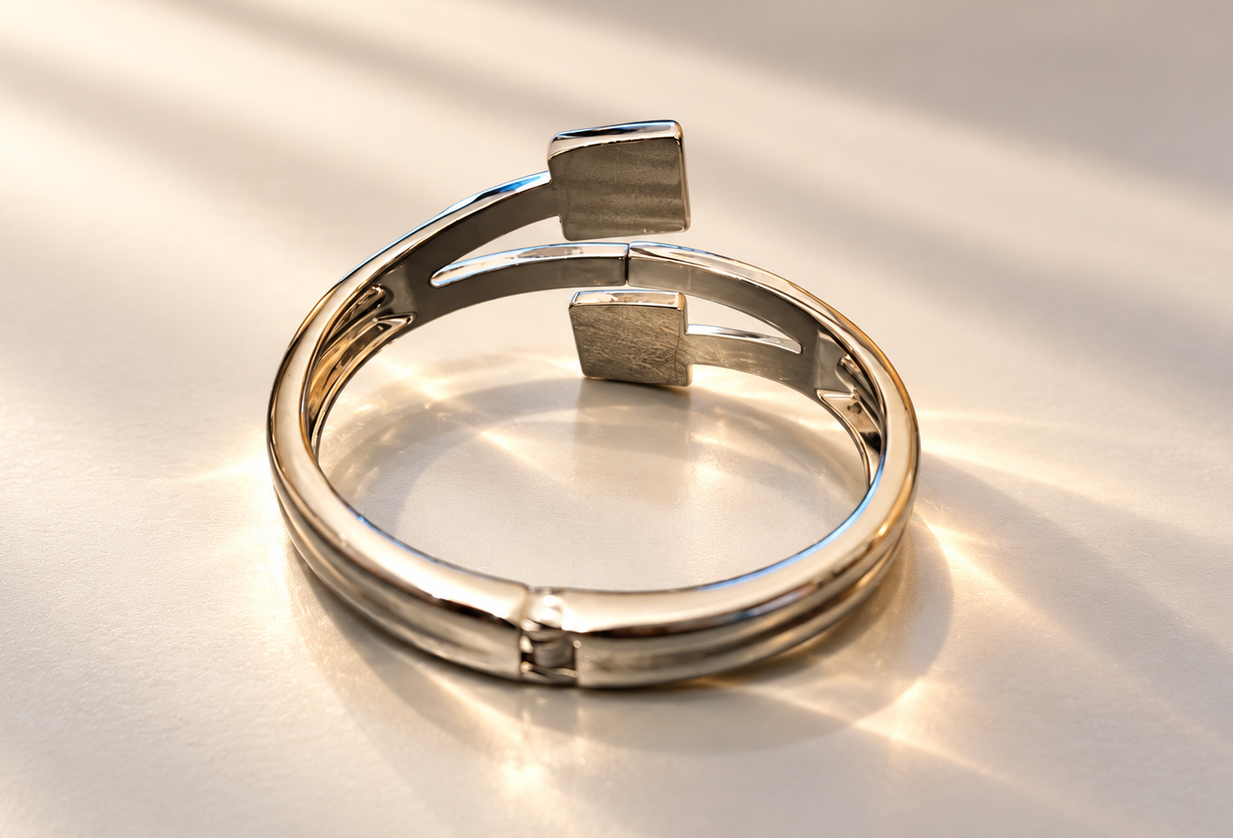 Bracelet Équilibre Double Carré Argenté — Bracelet rigide posé de trois-quarts sur un fond clair, lignes courbes ajourées, deux motifs carrés décalés et articulation visible au premier plan.