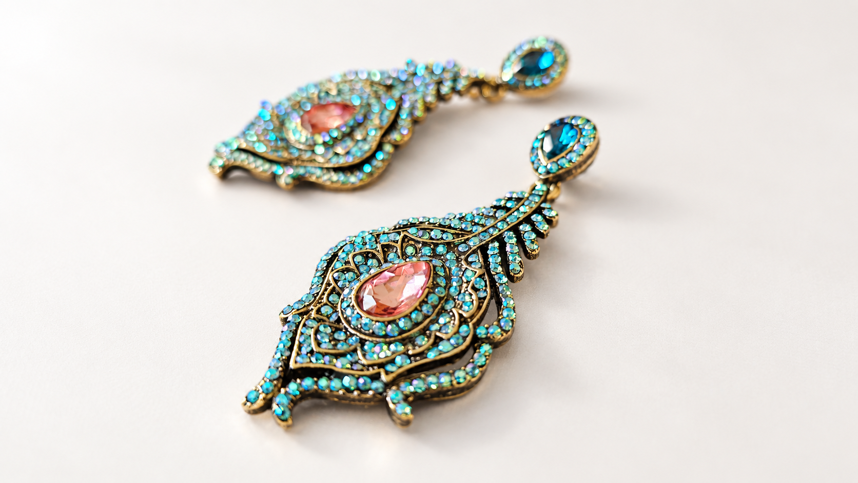 Boucles d’oreilles Parade Plumes Bleues & Roses — Paire vue en angle rapproché, une plume nette au premier plan montrant le relief doré, la goutte rose facettée et les reflets irisés bleu-vert.