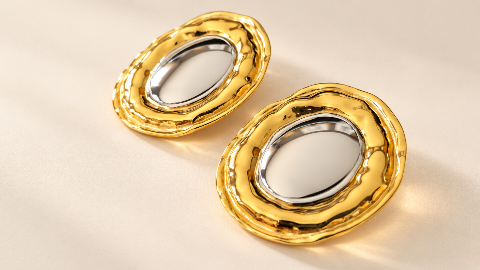 Boucles d’oreilles Halo Ovale Bicolore — Paire de boucles ovales disposées en diagonale sur un fond clair, avec larges contours dorés ondulés et bordures intérieures argentées autour des ouvertures.