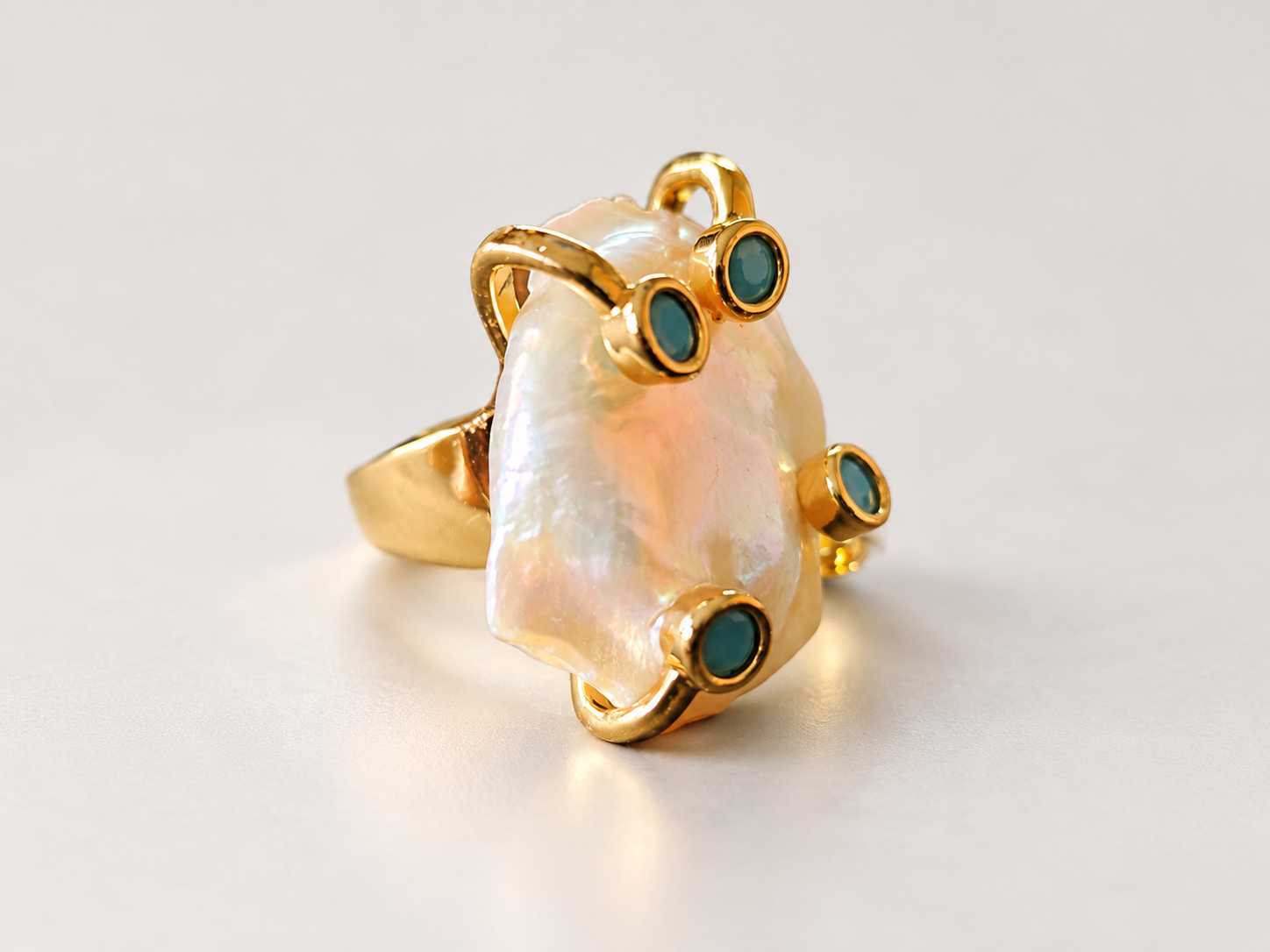 Bague Aurore Irisée aux Cabochons Bleu-Vert — Bague de trois-quarts avec anneau visible à gauche, élément central à reflets rosés et quatre cabochons bleu-vert bordés de doré.