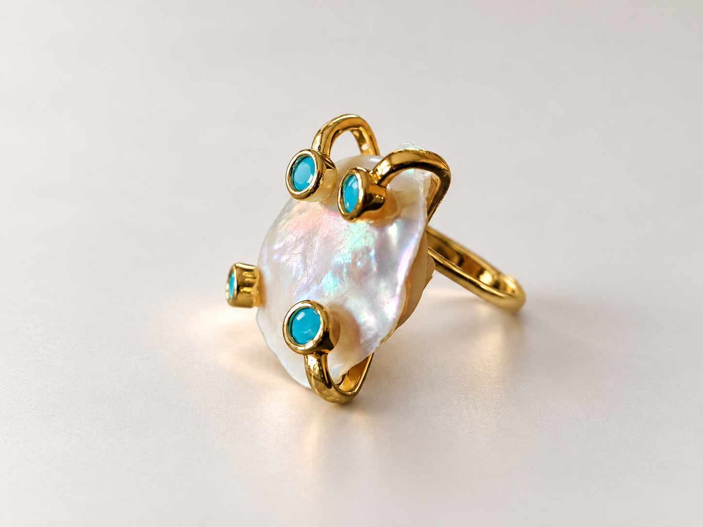 Bague Aurore Irisée aux Cabochons Bleu-Vert — Bague inclinée de trois-quarts, anneau à droite et courbes dorées en relief autour d’un motif irisé ponctué de cabochons bleu-vert.