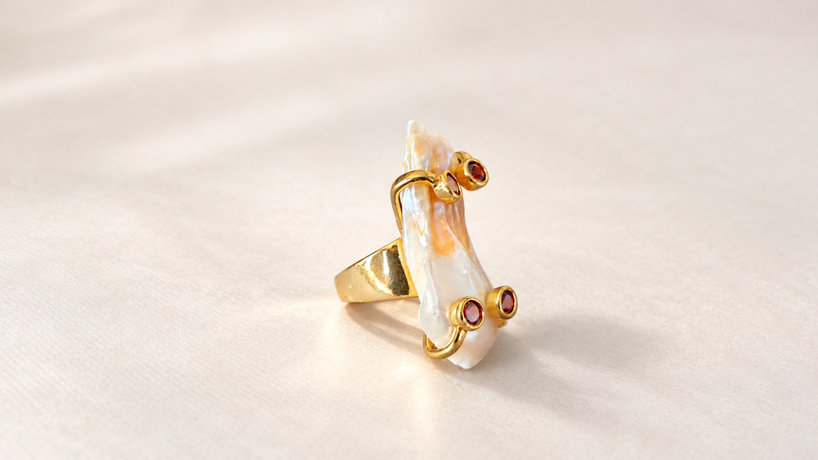 Bague Aurore Baroque aux Accents Rouges — Bague vue de côté, avec motif allongé en relief, attaches dorées courbes et éléments rouges ronds dépassant de la surface claire.